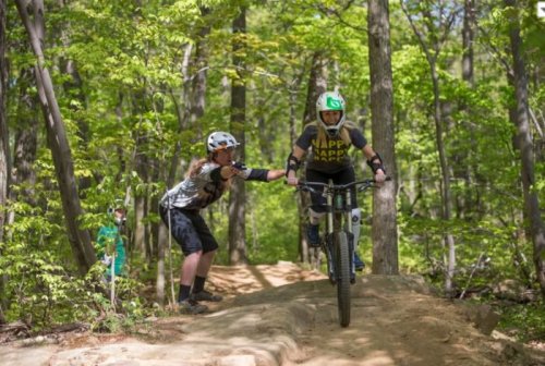 A mountain biker jumps off a dirt ramp while a friend assists from the side in a lush, green forest.