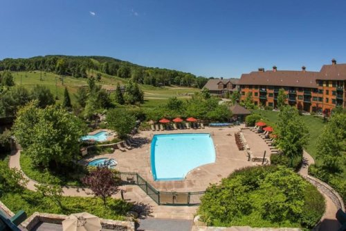 Aerial view of a resort pool area surrounded by greenery, with lounge chairs and hot tubs under a clear blue sky.