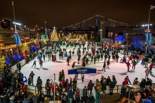 A bustling ice skating rink at night, surrounded by festive lights and a bridge in the background.