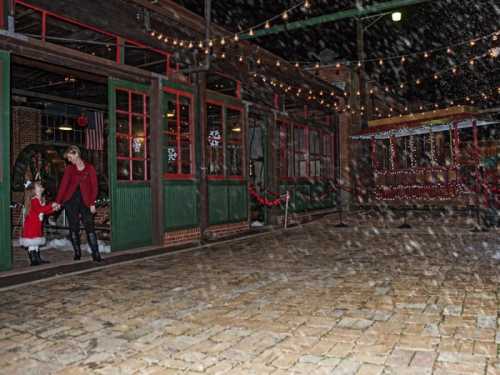 A woman and a child in festive attire walk together in a snowy, decorated outdoor area with string lights.