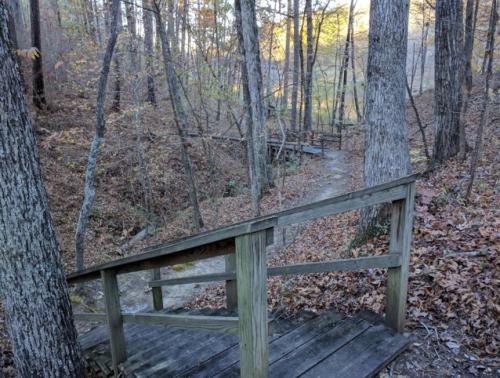 A wooden staircase leads down through a forested area with autumn leaves and trees, towards a path and bridge.
