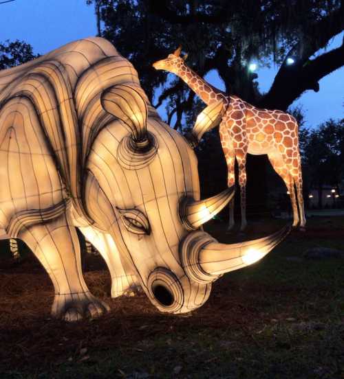 Illuminated sculptures of an elephant and a giraffe in a park at dusk, surrounded by trees.