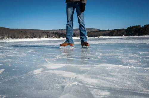 A person stands on a frozen lake, wearing jeans and brown boots, with a clear blue sky and trees in the background.