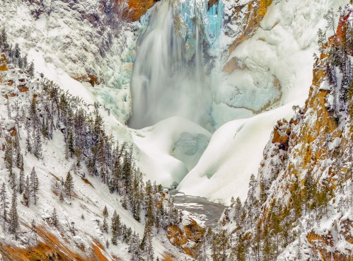 A frozen waterfall surrounded by snow-covered cliffs and evergreen trees in a winter landscape.