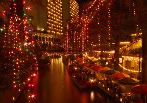 A festive riverside scene with colorful lights, umbrellas, and people dining along a beautifully illuminated canal.