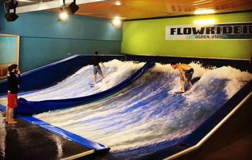 Two surfers ride artificial waves on a FlowRider attraction, with a spectator watching nearby.