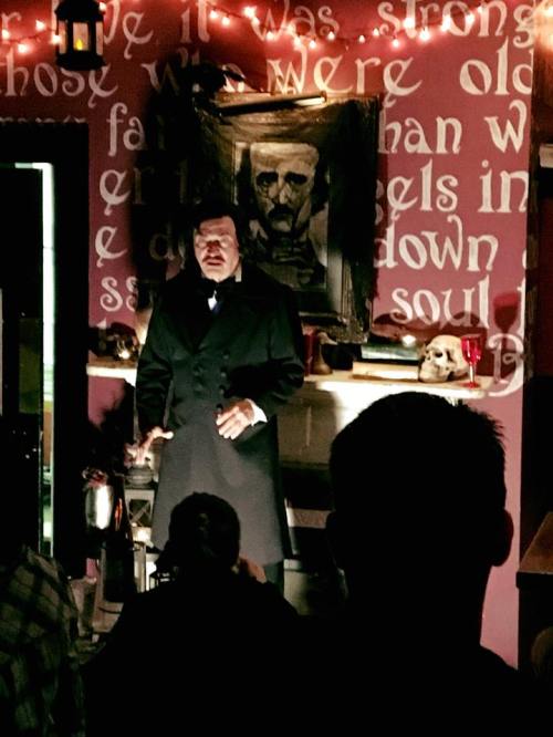 A performer in a dark coat stands in front of a portrait, speaking to an audience in a dimly lit room.