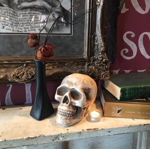 A decorative skull beside a black vase with dried roses, books, and a candle on a rustic table against a patterned wall.