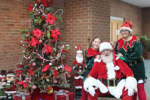 A cheerful Santa Claus poses with two women in festive outfits beside a decorated Christmas tree and holiday decorations.