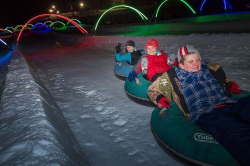 Three children enjoy snow tubing at night, surrounded by colorful lights and snow-covered hills.