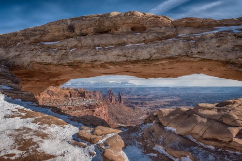 A stunning view through a natural rock arch, overlooking a vast desert landscape with snow-dusted ground.