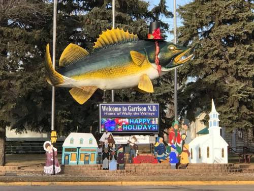 A large fish statue wearing a hat, with a holiday sign and festive decorations in a small town setting.