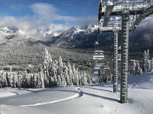 Snow-covered mountains and trees under a blue sky, with ski lift chairs and fresh tracks in the snow.
