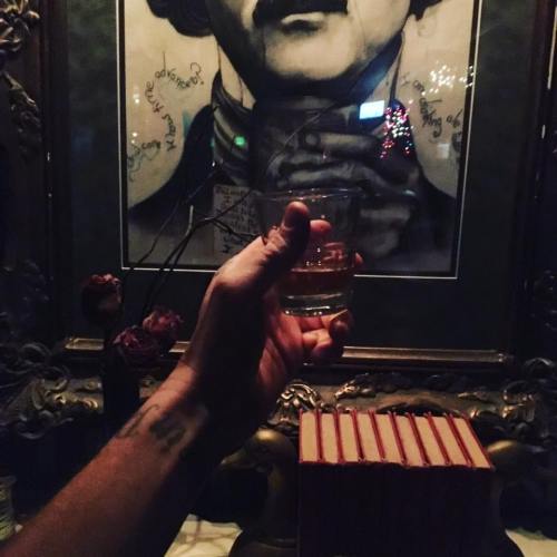 A hand holding a glass in front of a vintage portrait and a stack of books, with dim lighting and decorative elements.