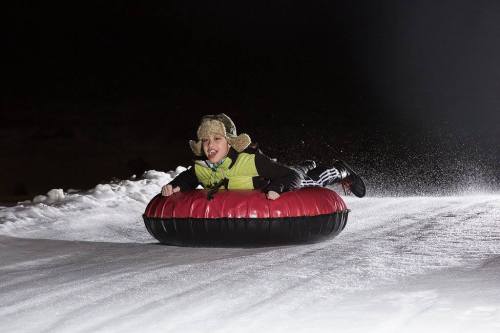 A child in a winter outfit joyfully sleds down a snowy hill on a red and black inflatable tube.