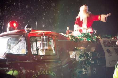 Santa Claus waves from a decorated fire truck during a festive parade, with snow falling around him.