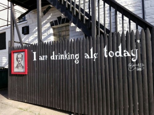A black wooden fence with a sign reading "I am drinking ale today" and a framed portrait above it.
