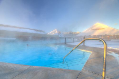 A misty outdoor pool surrounded by snow-covered mountains, with a metal railing leading into the water.