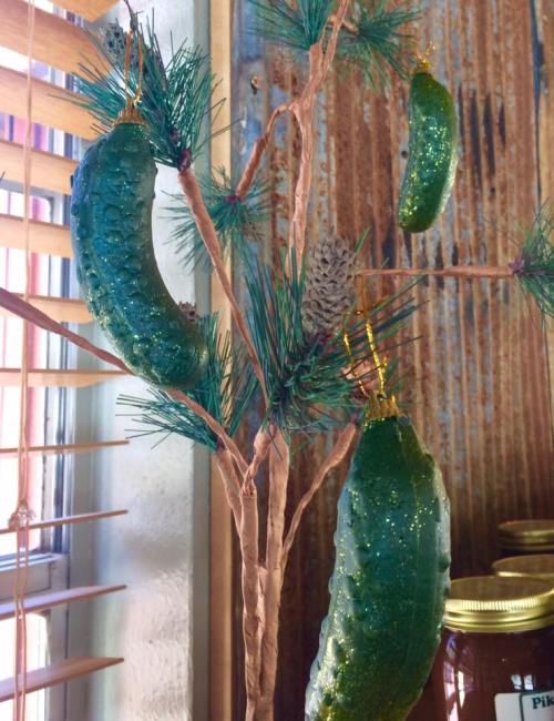 A small tree decorated with shiny green pickle ornaments and a pinecone, set against a rustic background.