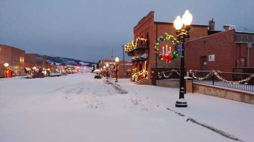 Snow-covered street decorated with holiday lights and wreaths, lined with buildings and lampposts in a quiet town.