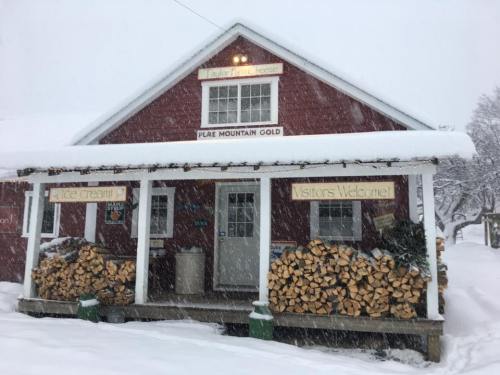A red building with a snowy porch, stacked firewood, and a sign welcoming visitors, set in a winter landscape.