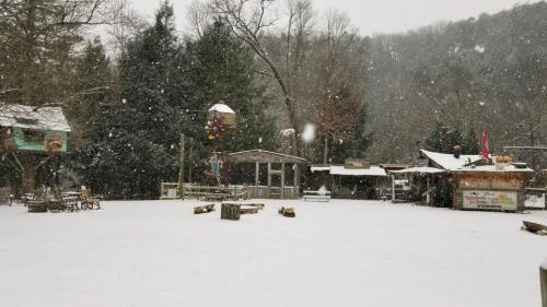 A snowy outdoor scene with a rustic area, featuring wooden structures and trees in the background. Snowflakes are falling.