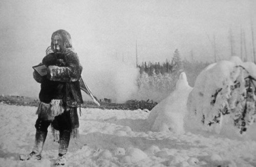 A person stands in a snowy landscape, holding a book, with trees and a snow-covered mound in the background.