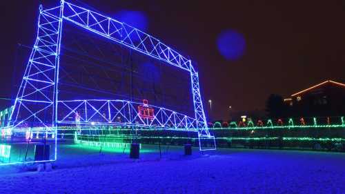 A large illuminated structure with blue and green lights in a snowy park at night.