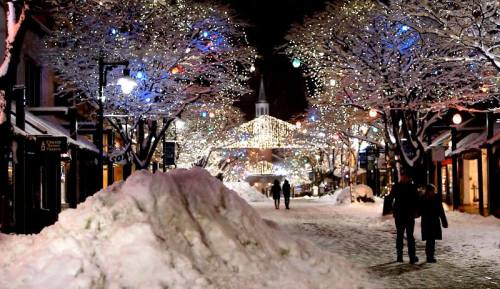 A snowy street at night, lined with trees adorned in colorful holiday lights, creating a festive atmosphere.