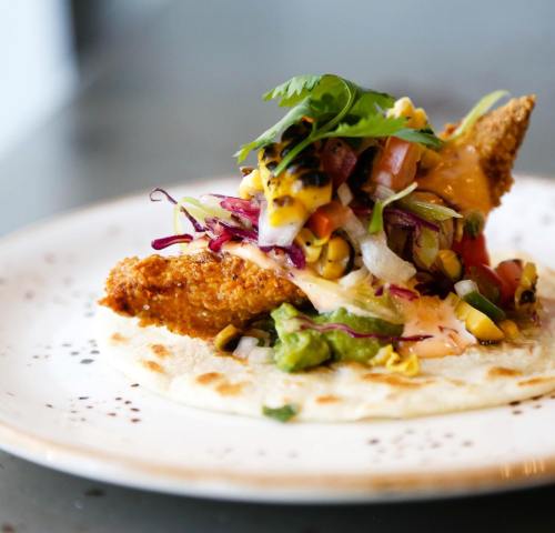 A crispy fried taco topped with colorful salsa, guacamole, and fresh cilantro on a plate.