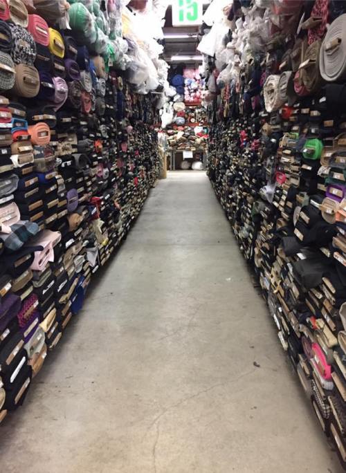 A narrow aisle between tall shelves filled with various rolls of fabric and ribbons in a craft store.