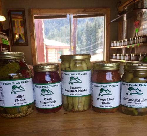 Jars of pickles and salsas on a wooden shelf, labeled with "Pikes Peak Pickles" in a rustic setting.