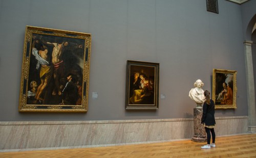 A visitor admires paintings and a bust in an art gallery, featuring dramatic lighting and classic artwork.