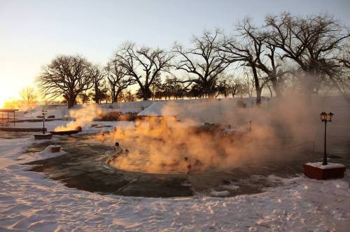 Steam rises from a hot spring surrounded by snow, with trees silhouetted against a sunset in the background.