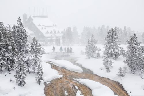 A snowy landscape with a river and a group of people walking on a bridge, surrounded by trees and a distant lodge.