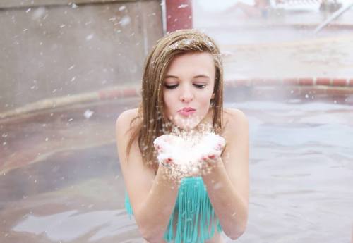 A young woman in a turquoise bikini blows a cloud of powder into the air while standing in a hot spring.