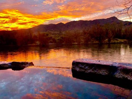 A serene sunset over a river, with vibrant orange and purple hues reflecting on the water and mountains in the background.