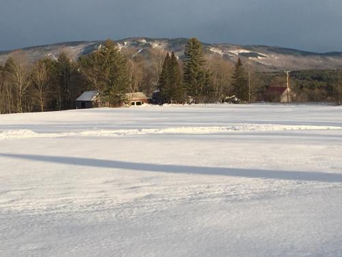 Snow-covered landscape with distant mountains and trees, featuring a few buildings in the foreground.