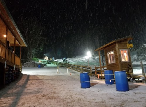 A snowy night scene at a ski resort, featuring a wooden lodge, a ticket booth, and illuminated ski slopes in the background.