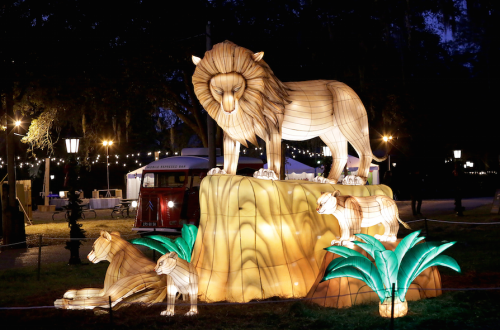 A glowing lion sculpture stands on a rock, surrounded by smaller lions and illuminated greenery at night.