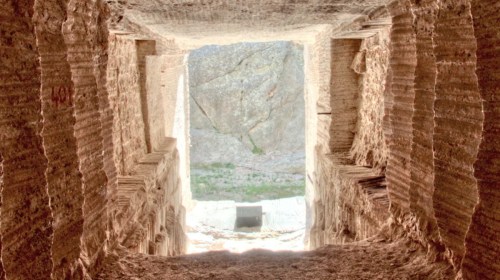 View of a stone tunnel leading to a bright opening, with textured walls and a rocky landscape visible outside.