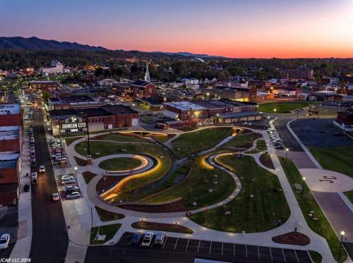 Aerial view of a park with winding paths and lights, surrounded by a small town at sunset with mountains in the background.
