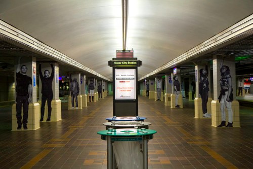 Empty subway station with tall pillars featuring large black-and-white portraits, and a central information stand.