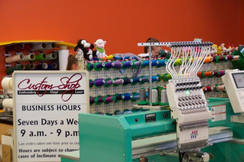 A colorful embroidery machine in a shop with spools of thread and a sign detailing business hours.