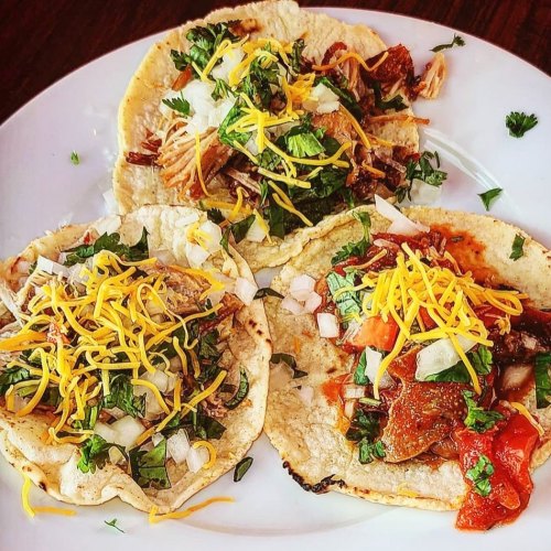 Three tacos on a plate, filled with shredded meat, onions, cilantro, and topped with cheese.