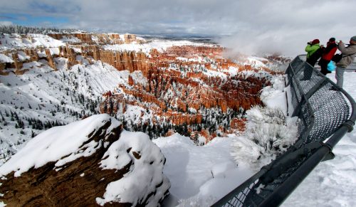 Snow-covered landscape of Bryce Canyon with visitors at a viewpoint overlooking the stunning red rock formations.