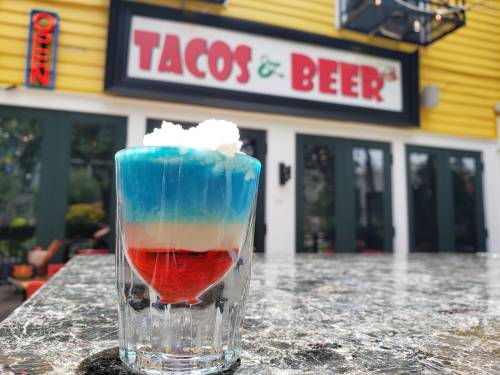 A layered dessert in a glass with blue, white, and red colors, set against a restaurant backdrop that reads "Tacos & Beer."