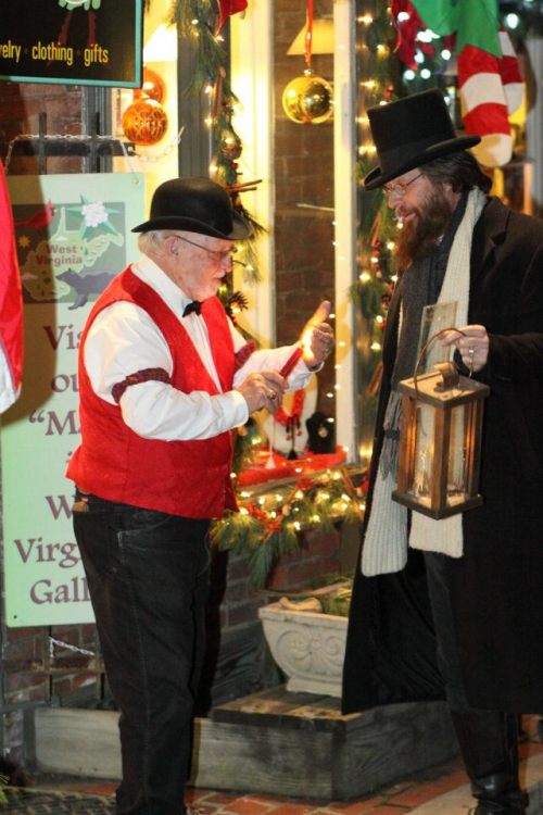 Two men in festive attire, one with a lantern, stand outside a decorated shop, surrounded by holiday decorations.