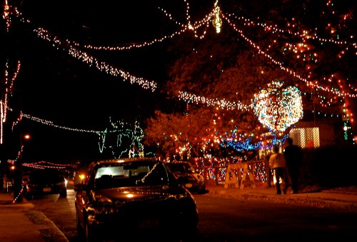 A street decorated with colorful holiday lights at night, with cars parked and people walking nearby.