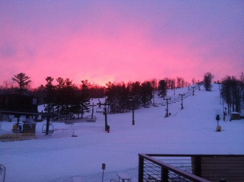 A snowy ski slope at sunset, with pink and purple skies and silhouetted trees in the background.